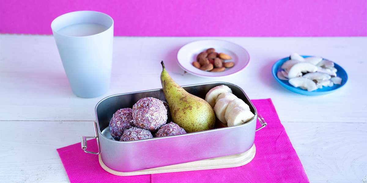 Energy Balls Himbeere in einer Lunchbox zusammen mit einer frischen Birne und Bananenscheiben. Darunter liegt eine bunte Serviette. Links steht ein Glas Milch und rechts zwei kleine Teller mit Kokosnusschips und Mandeln