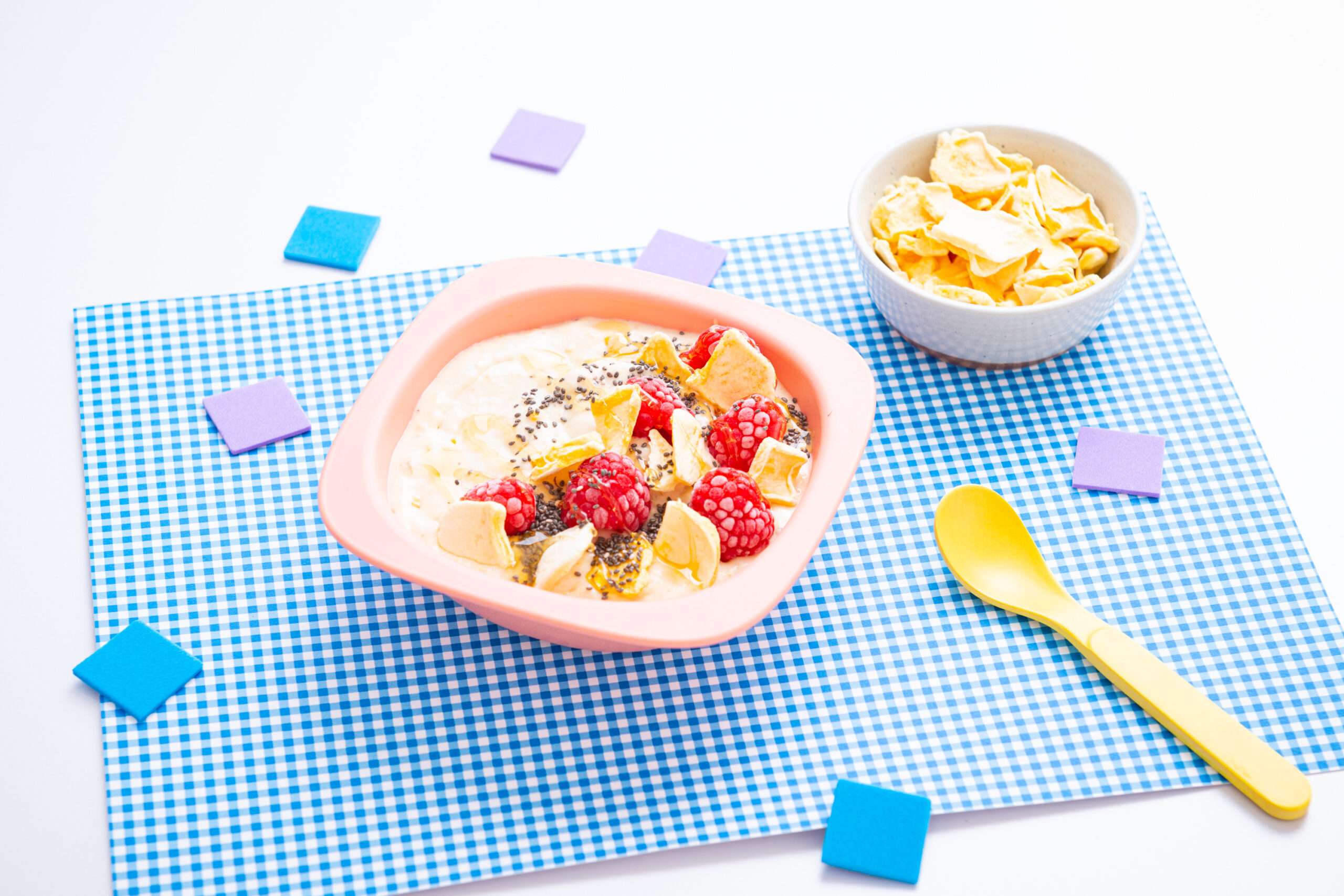 Mango Nicecream in einer Schüssel angerichtet mit Mango Fruchtchips, Himbeeren und Chia Samen. darunter liegt ein Platzdeckchen. Ein Löffel und ein Schüssel mit Fruchtchips stehen dabei.