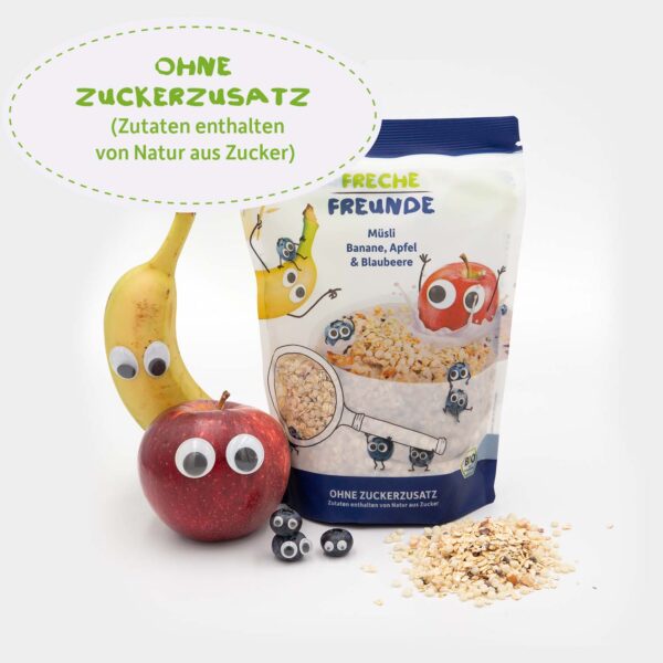 Die Verpackung des Freche Freunde Bio Müslis steht zwischen einer echten Banane, einem Apfel und drei Blaubeeren – alle mit frechen Wackelaugen versehen. Vor der Packung liegt Müsli verstreut. Text in Sprechblase: „Ohne Zuckerzusatz (Zutaten enthalten von Natur aus Zucker)“.