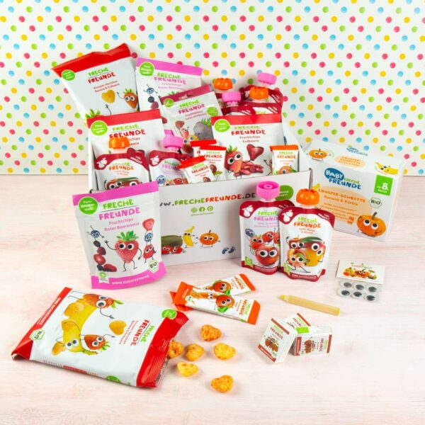 Das Freche Freunde Bio Probierpaket in geöffneter Versandbox – randvoll mit knusprigen, fruchtigen und quetschigen Snacks für kleine Snack-Fans. Bunte Punktetapete im Hintergrund, einige Produkte stehen locker davor.