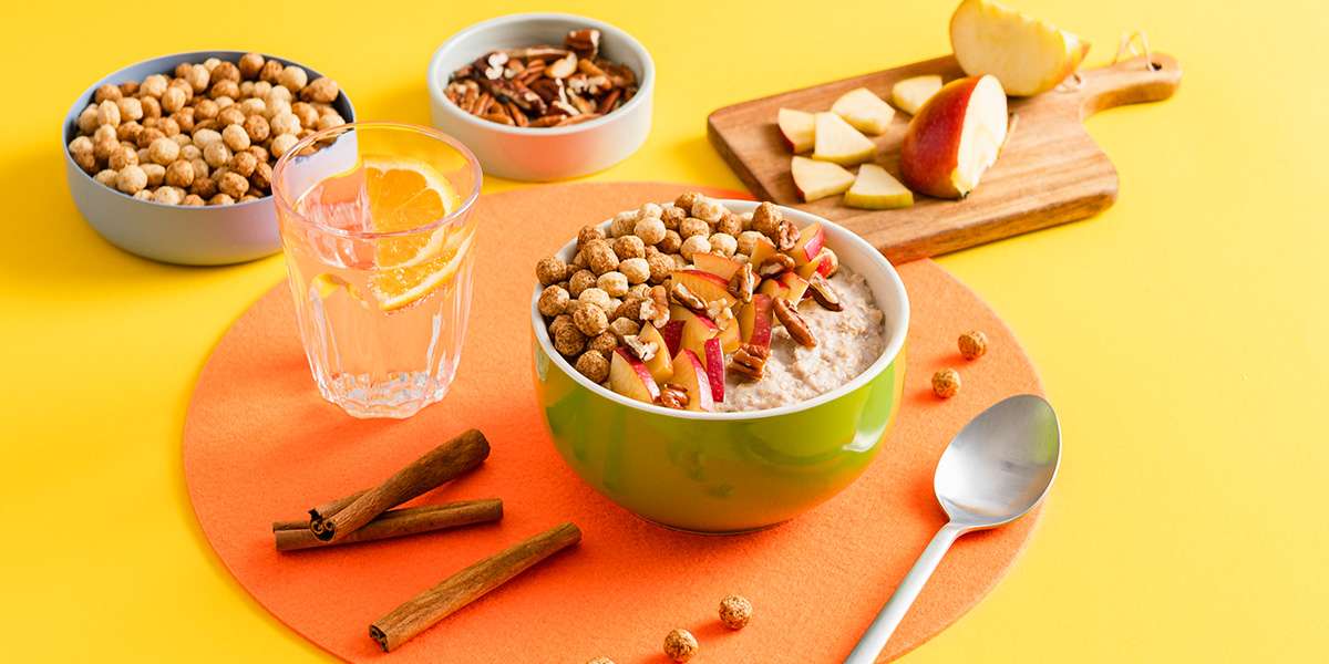 Apfel Zimt Porridge in einer Schüssel serviert. Frische Apfelspalten und Cerealien als Topping. Daneben steht ein Glas Wasser und weitere Schüsseln gefüllt mit Pekan Nüssen und Cerealien.