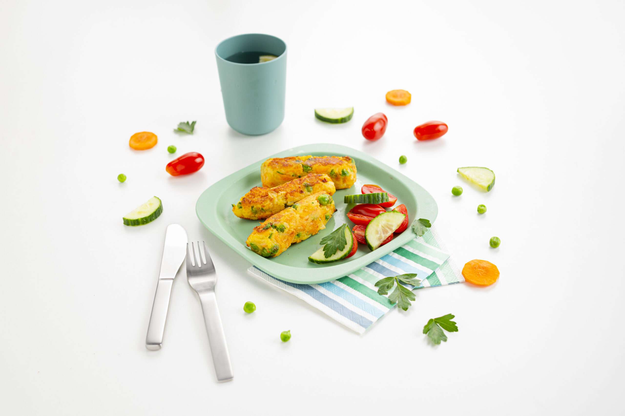 Gemüsestäbchen auf einem Teller serviert zusammen mit frischen Gemüse. Der Teller liegt auf einer Serviette. Dazu ein Becher mit Schorle.