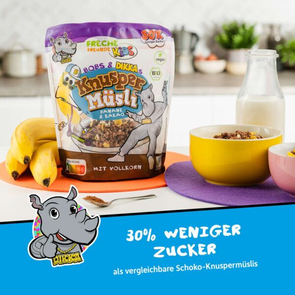 Freche Freunde Bio Bobs & DIKKAs Knusper Müsli Banane & Kakao in einer Frühstücksszene mit frischer Banane, Milch und bunten Schalen. Die Packung steht auf einem orangen Untersetzer, daneben eine gelbe und rosa Müsli-Schale – bereit für ein freches Frühstück mit 30 % weniger Zucker.