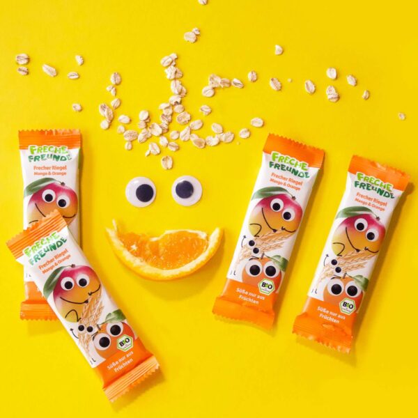 Vier freche Riegel Mango & Orange tanzen rund um eine Orangenspalte mit Kulleraugen und aufgestreuten Haferflocken – das fröhliche Gesicht bringt alle zum Schmunzeln.