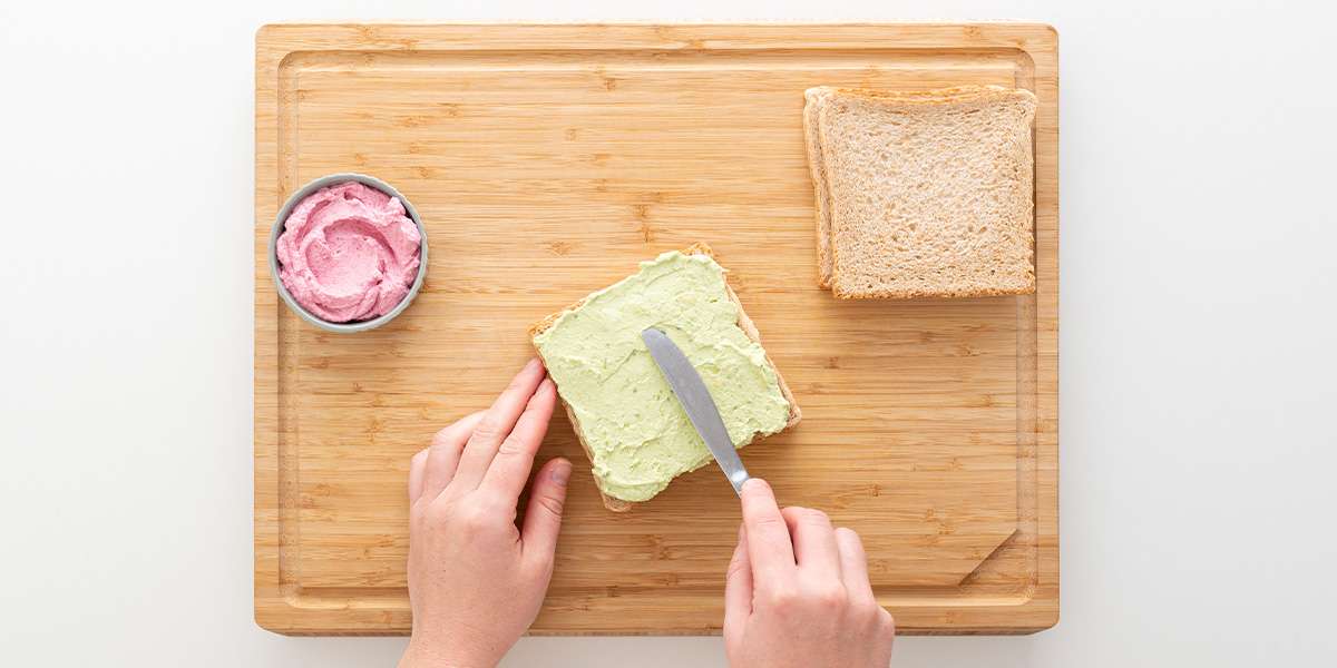 Das Gemüse-Sandwich wird mit der Avocado-Creme mit einem Messer geschmiert. Daneben ist die rosa Rote Bete Creme in einer Schüssel und 2 weitere Scheiben Brot.