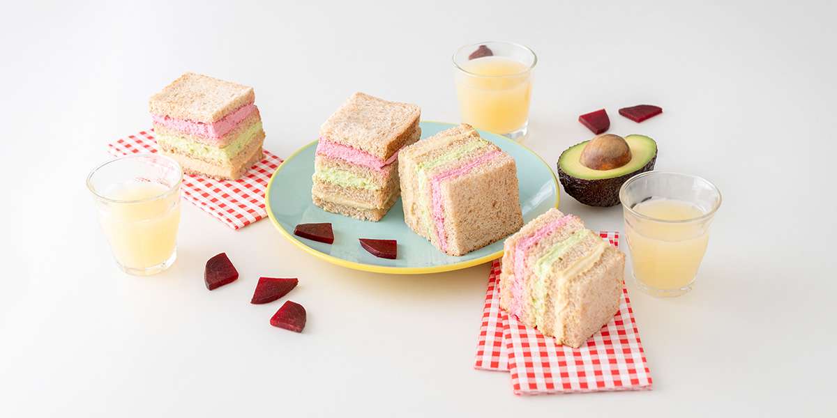 Gemüse-Sandwich mit rosa, grünem & beigem Aufstrich auf einem Teller, daneben Rote Bete Stückchen und aufgeschnittene Avocado