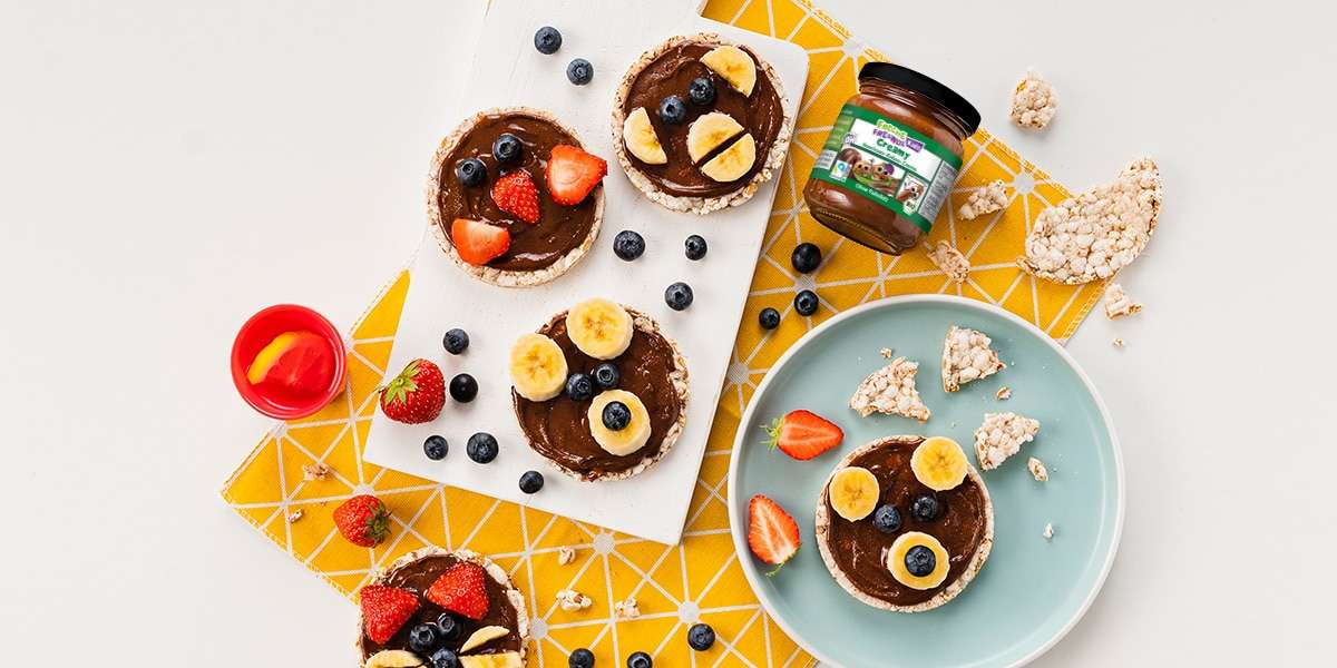 Reiswaffeln mit Haselnussmus bestrichen und mit Gesichtern aus Bananenscheiben, Blaubeeren und Erdbeeren dekoriert. Ein Glas Creamy Haselnuss-Kakao Creme liegt dabei.