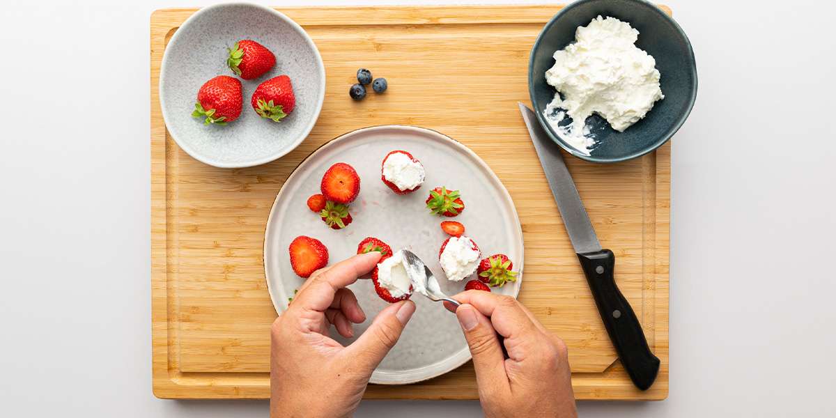 Für die Erdbeer Zwerge wird Quark auf die geteilten Erdbeeren gestrichen mit einem Löffel. Daneben die restlichen Erdbeeren, der Quark und ein Messer.