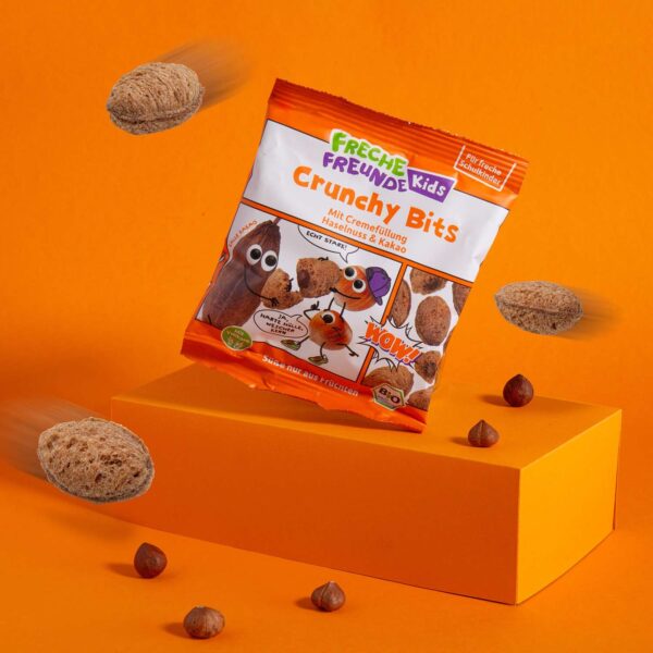 Freche Freunde Kids Bio Crunchy Bits auf einem orangem Podest und Hintergrund und rundherum fliegende Crunchy Bits.