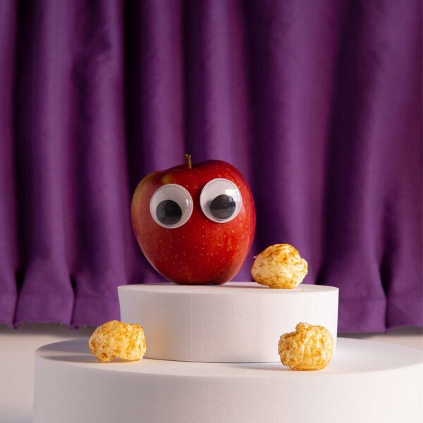 Freche Freunde Bio Alfreds Poppies Apfel Karamell Style auf einem kleines Podest vor einem lila Vorhang, daneben Alfred Apfel mit Wackelaugen.