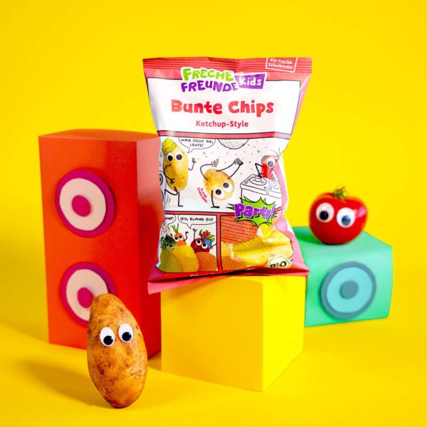 Freche Freunde Kids Bunte Chips Ketchup-Style Verpackung im Comic Design auf gelben Podest und Hintegrund, daneben Klaus Kartoffel und Tom Tomate mit Wackelaugen.