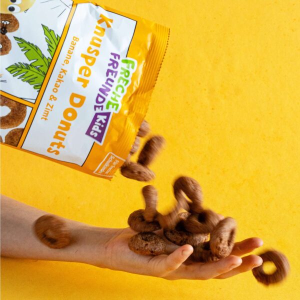 Freche Freunde Bio Knusper Donuts fliegen dynamisch aus der Packung Banane, Kakao & Zimt direkt in eine Kinderhand. Action pur mit frechem Snackspaß auf leuchtend gelbem Hintergrund – knusprig, wild und natürlich lecker!
