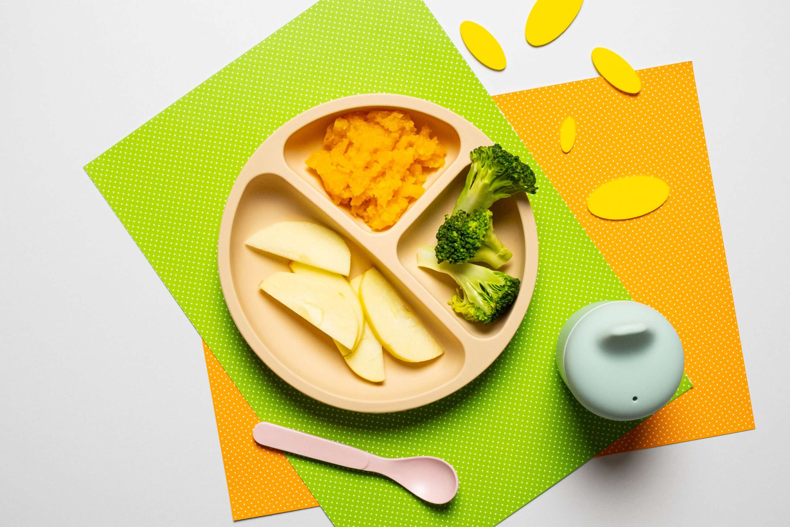 Kürbispüree für Babys mit Apfelspalten und Brokkoliröschen auf einem Menü Babyteller dazu ein Trinkbecher mit Wasser