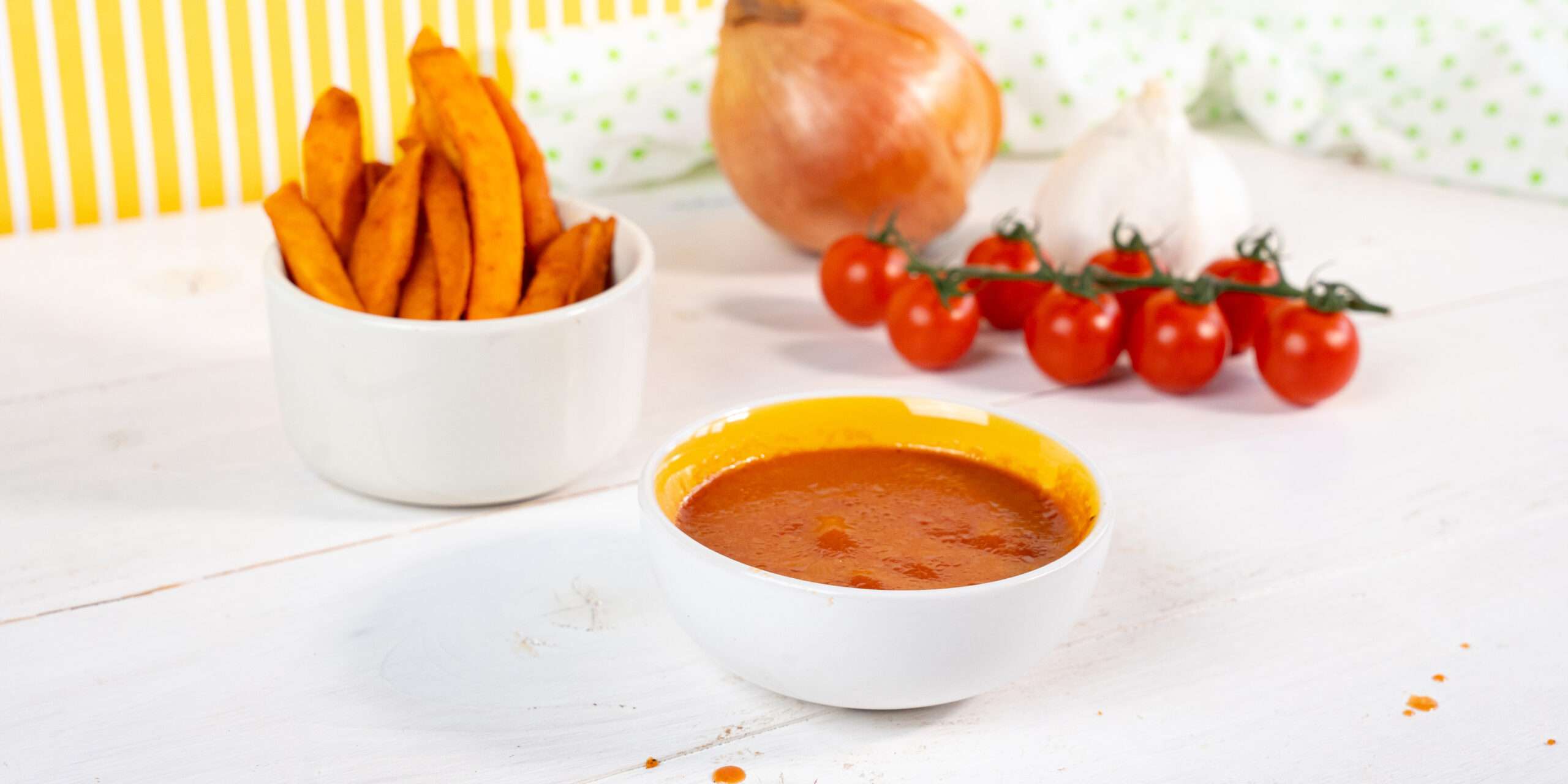 Ketschup ohne Zucker in einer Schüssel serviert. Dazu eine Schüssel mit Süßkartoffel-Pommes. Im Hintergrund liegen frische Tomaten & eine Zwiebel.