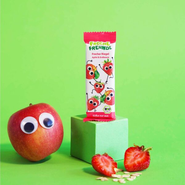 Freche Freunde Bio Frecher Riegel Apfel & Erdbeere posiert auf grünem Podest. Neben dem Riegel mit Alfred Apfel und Edda Erdbeere eine echte Erdbeere und ein Apfel mit Wackelaugen.