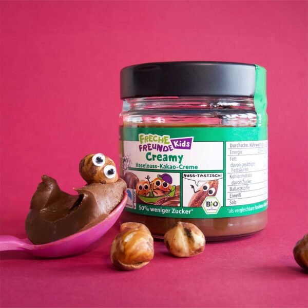 Freche Freunde Kids Bio Creamy Haselnuss-Kakao-Creme im Glas auf pinkem Hintergrund, davor ein Löffel voll cremiger Schoko-Nuss-Aufstrich mit zwei kichernden Hassan Haselnüssen als Figur. Ein weiterer frecher Haselnuss-Charakter beobachtet das Schokospektakel aus dem Hintergrund.