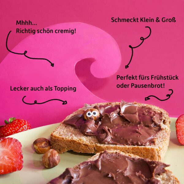 Zwei Scheiben Brot mit der Freche Freunde Kids Bio Haselnuss-Kakao-Creme bestrichen, dekoriert mit Erdbeeren und einem frechen Haselnuss-Charakter, der aus der Creme herauslugt. Text auf dem Bild: „Mhmm… richtig schön cremig!“, „Perfekt fürs Frühstück oder Pausenbrot!“, „Lecker auch als Topping“, „Schmeckt Klein & Groß“.