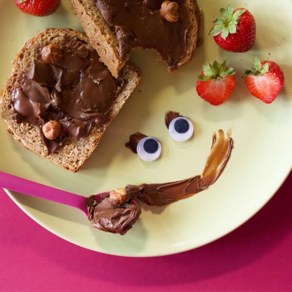 Lustig angerichteter Teller mit zwei Scheiben Brot mit Freche Freunde Kids Bio Creamy Haselnuss-Kakao-Creme, frischen Erdbeeren und einem frechen Schoko-Gesicht aus Creme, Wackelaugen und einem Löffel mit Hassan Haselnuss.