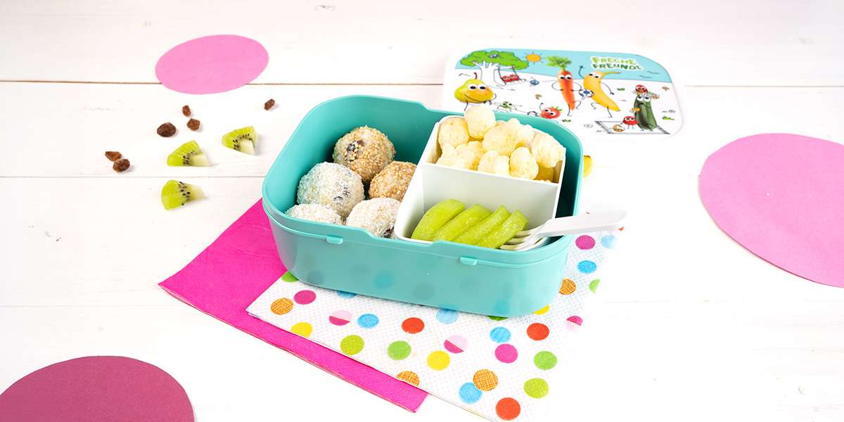 Freche Freunde Brotdose gefüllt mit Mandel Kokos Bällchen, Kiwi Scheiben und Party Pops Popcorn. Darunter liegen bunte Servietten. Der Deckel der Brotdose ist mit der Freche Freunde Welt bedruckt.