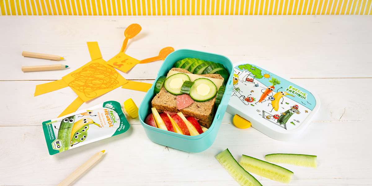 Lustiges Eulen Sandwich in der Freche Freunde Brotdose. Gefüllt mit Gurkenscheiben und Apfelspalten. Daneben liegt ein Quetschie, Buntstifte und eine Kinderzeichung.