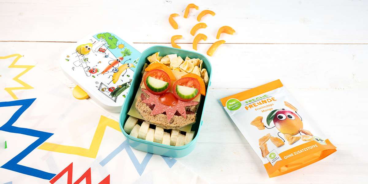 Ein Monster Toast in der Freche Freunde Brotdose. Die Augen sind Tomatenscheiben mit Gurkenhälften und eine Salami im Zickzack Schnitt sind die Zähne. Daneben liegt eine Packung Mango Fruchtchips.