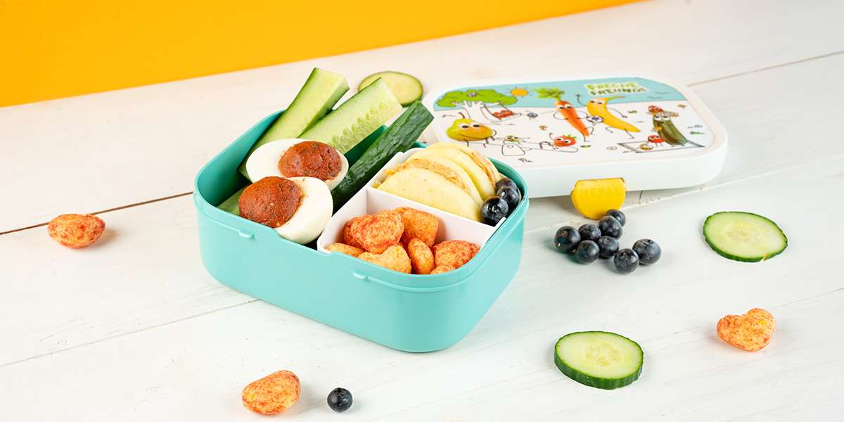 Freche Freunde Brotdose gefüllt mit Apfelringe mit Erdbnussbutter, gefülltem Ei, Gurkensticks, Blaubeeren und Knusper Herzchen. Dahinter liegt der Deckel der Lunchbox.