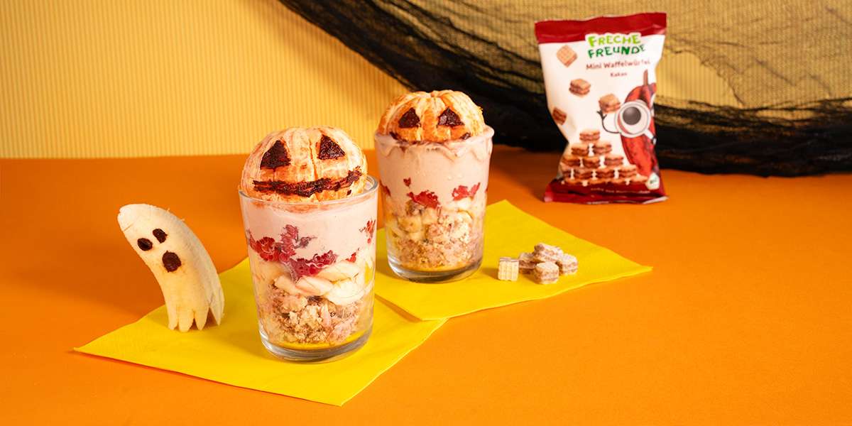 Zwei gruselige Halloween Desserts für Kinder sind im Glas angerichtet. Eine Mandarine mit Halloween Gesicht liegt oben auf dem Dessert auf. Daneben eine halbe Banane mit Gesicht als Geist. Im Hintergrund stehen die Mini Waffelwürfel welche im Rezept verwendet werden.