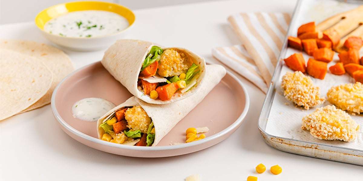 Zwei leckere Crispy Nuggets Wraps serviert auf einem Teller. Daneben liegt das Blech mit Chicken Nuggets und Süßkartoffeln aus dem Ofen und eine eine Schüssel mit einen Dip.