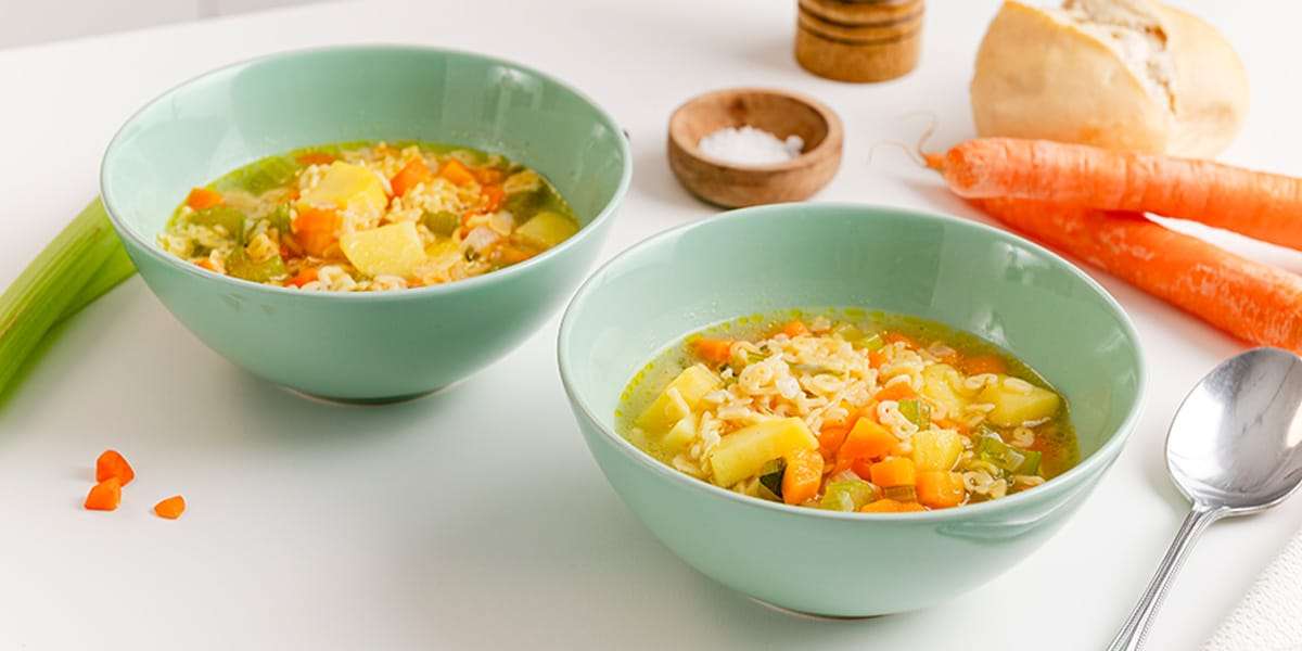 Zwei Suppenschüsseln gefüllt mit Nudelsuppe und Gemüse. Im Hintergrund liegen frische Karotten und ein Brötchen.