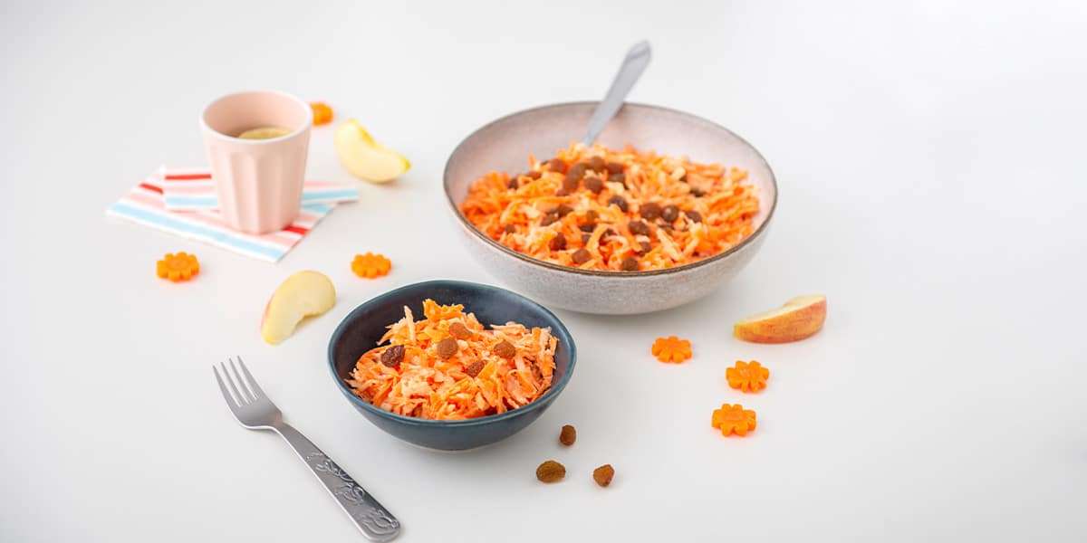 Karotten Apfel Salat in einer Schüssel serviert, daneben steht eine kleine Schüssel mit Salat & einer Gabel sowie ein Becher mit Wasser